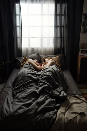 Couple sleeping peacefully under warm blankets in cozy bedroom with soft natural light streaming through window creating serene atmosphereの写真素材