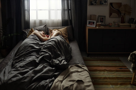Woman lying asleep under a blanket in a dimly-lit cozy bedroom with various decorations on bedside table and wallの写真素材