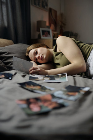 Woman lying on bed, holding photograph, appearing in deep thought surrounded by scattered photographs, creating nostalgic ambiance. Bedroom setting with decor visible in backgroundの写真素材