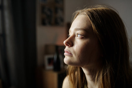 Woman with long hair sitting indoors, looking out of window while thinking deeply. Sunlight casting...