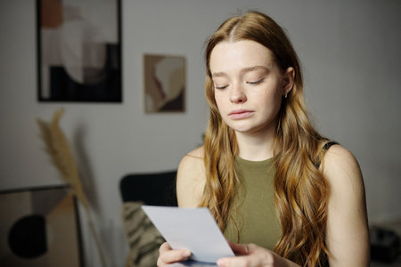 Young woman with long hair is holding and reading letter in modern living room with abstract wall art and dried plant decoration, showing thoughtful expressionの写真素材