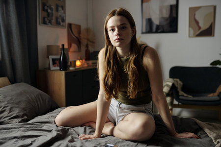 Portrait of young woman sitting on bed in cozy room with artistic decorations and dim lighting creating a serene atmosphereの写真素材