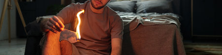Portrait of man sitting in dimly lit bedroom holding burning paper while wearing casual clothes. The background features a neatly made bed, and dark wallsの写真素材