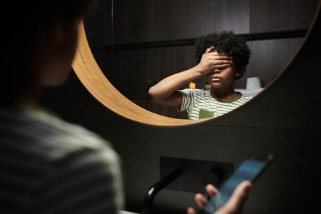 Young black boy standing in front of mirror covering eyes with hand while holding smartphone, getting messagesの写真素材