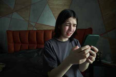 Young girl with dark hair sitting on bed, holding smartphone with confused expression, illuminated...