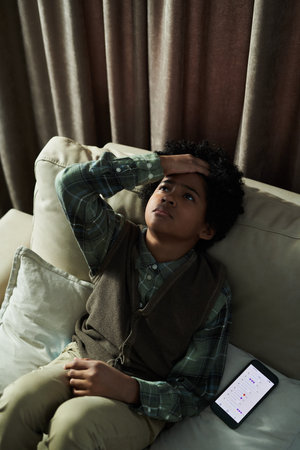 Young boy reflecting with hand on head sitting on couch next to mobile phone with open app on screen