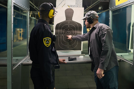 Two men at shooting range, one watching other aiming at target silhouette with firearm. Strong focus...