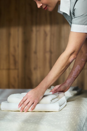 Person arranging neatly folded towels on bed in cozy spa environment with wooden background showing relaxation and care for customer needsの写真素材