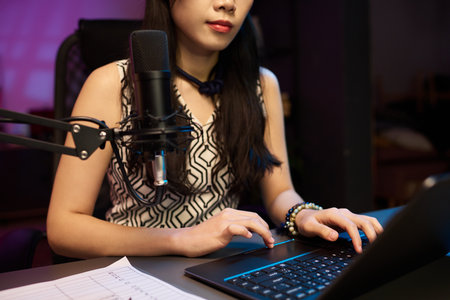 Woman sitting at desk, using a laptop and microphone during podcast recording. Capturing a moment of focus and engagement in a creative environmentの写真素材