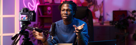 Portrait of passionate Black person sitting in front of camera, recording engaging content in a studio setting with neon lights in the backgroundの写真素材