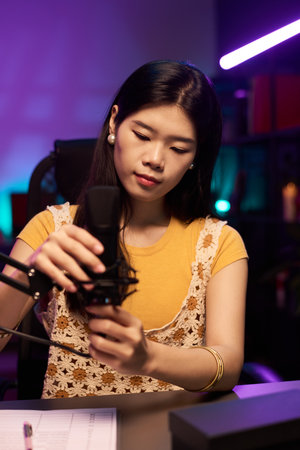 Asian girl adjusting microphone in a dimly lit studio with purple accents, preparing for recording session. Focused and attentive, she wears a yellow top and lace vest, holding microphone standの写真素材