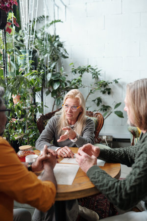 Three people engaged in animated discussion at a cozy indoor cafe with plants and warm lighting, one elderly person wearing glasses focused on communicating their ideasの写真素材