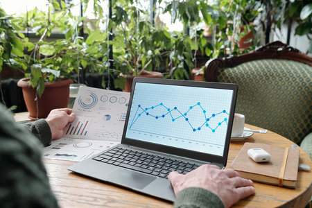 Hands of adult person analyzing financial charts on laptop surrounded by plants. Office setup...