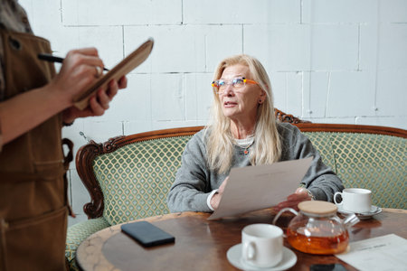 Elderly woman holding document during meeting in cozy cafe, engaging in conversation with another...