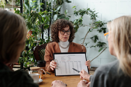 Woman holding a financial report while discussing with two other people during a business meeting surrounded by indoor plants and wooden furnitureの写真素材