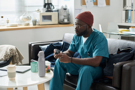 Wearing teal scrubs and a red beanie, an individual using a smartphone in a modern office with various devices and files organized around them creating a productive atmosphereの写真素材