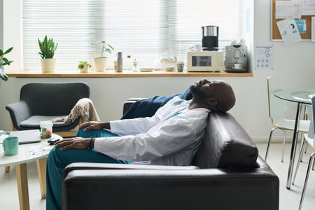Wearing the white coat, doctor is sitting on couch with closed eyes. Office setting includes chairs, table with devices, plants, and medical equipmentの写真素材