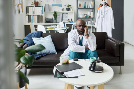 Tired doctor sitting in hospital lounge, resting head on hand, surrounded by documents, coffee mug, and medical equipmentの写真素材
