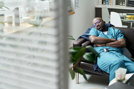 Tired medical professional sleeping in clinic break room, wearing scrubs, with medical equipment visible in background. Scene captures exhaustion of healthcare workersの写真素材