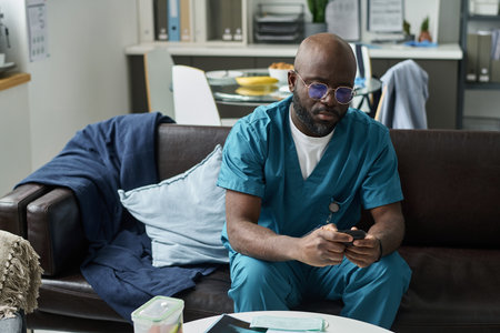 African American male nurse sitting in hospital lounge, wearing scrubs and spectacles, looking focused and contemplative, ready to assist patients and perform healthcare dutiesの写真素材