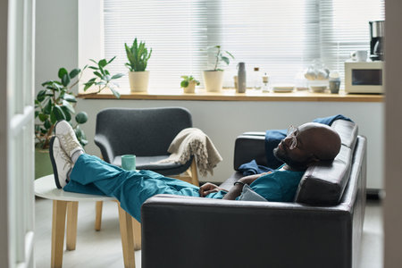 Medical professional resting in break room, lying on a couch with closed eyes. Green plants and...