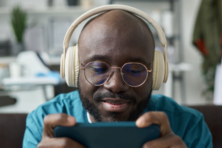 Close-up of Black person wearing headphones and glasses, engrossed in using smartphone. Seated...