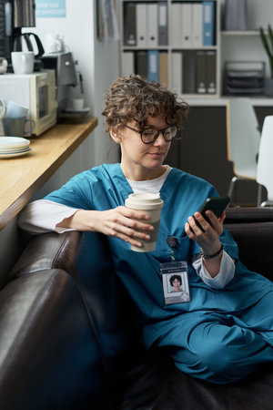 Medical professional wearing scrubs and glasses relaxing with coffee and smartphone in hospital...