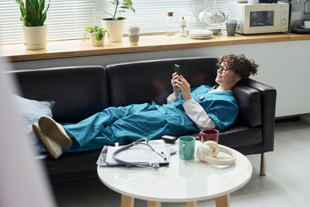 Medical professional in scrubs reclining on couch in break room, looking at phone and relaxing....
