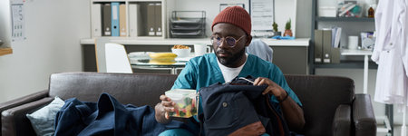 Man wearing medical scrubs and red beanie packing lunch in clear container while sitting on couch in office setting with shelves and files in backgroundの写真素材