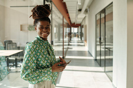 Black woman smiling and holding smartphone while standing in modern coworking office hallway with glass wallsの写真素材