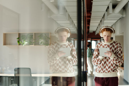 Young Caucasian man with headphones standing in modern coworking space using digital tablet,...
