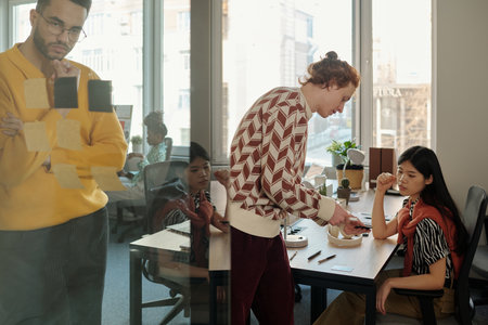 Man standing by glass wall writing on sticky notes while young adult Caucasian man and Asian woman...