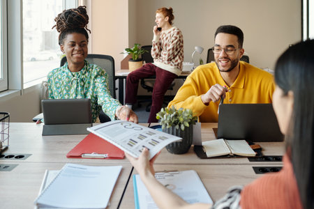 Young Black woman and man collaborating at coworking table with digital tablets while Caucasian man talking on phone, Asian woman handing documentの写真素材
