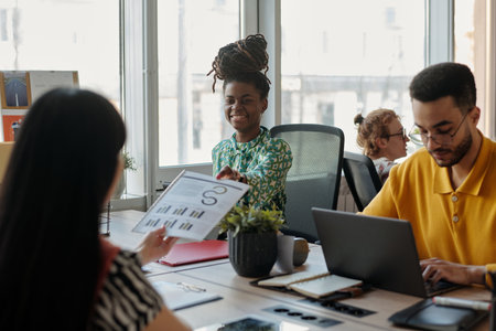 Young adult Black woman smiling while discussing business charts with colleague across desk, young adult man working on laptopの写真素材