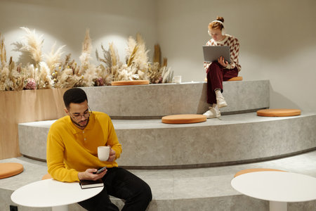 Black man sitting on tiered seating holding smartphone and coffee while Caucasian man with red hair...