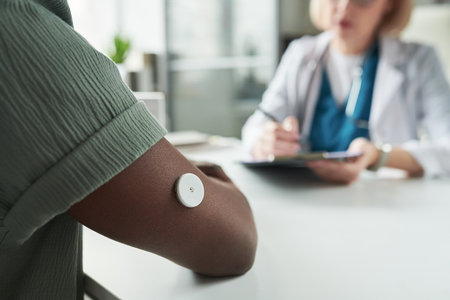 Black young adult man wearing glucose monitor on upper arm consulting with Caucasian middle aged female doctor in medical office, doctor writing notes during diabetes management appointmentの写真素材