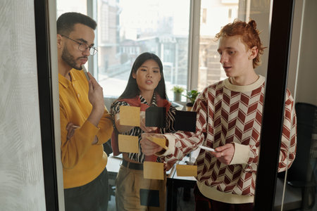 Caucasian man explaining project details using sticky notes on glass wall while young adult Black...