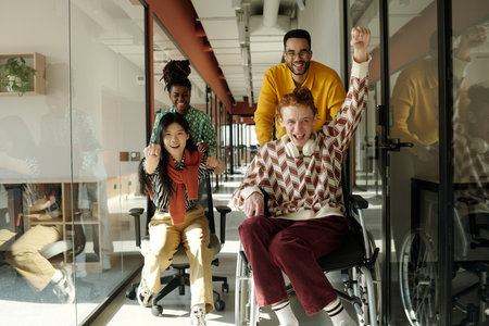 Diverse young adults in creative coworking space, Caucasian man in wheelchair being pushed by Black man while Asian and Black women cheer and smileの写真素材