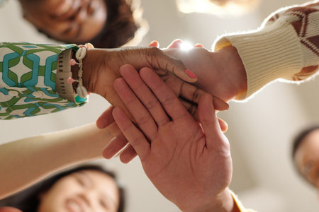 Diverse group of young adult men and women stacking hands in creative coworking space, showing close...