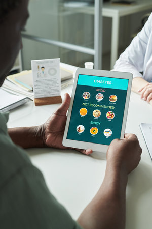 Middle aged Black man holding digital tablet displaying diabetes dietary recommendations during consultation with Caucasian female doctor in medical office, discussing healthy eating choicesの写真素材