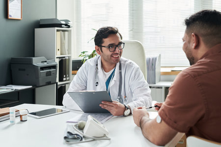 Middle aged Caucasian male doctor smiling while consulting middle aged Hispanic man with diabetes in medical office, patient sitting at desk with glucose monitor on arm, discussing health managementの写真素材