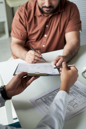 Middle aged Caucasian man consulting with doctor in medical office, holding pen and signing medical...