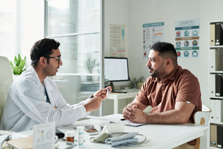 Middle aged Caucasian man sitting across from male doctor in medical office discussing diabetes management with glucose meter on desk, both focusing on healthcare consultationの写真素材