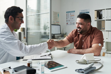 Middle aged Caucasian man with diabetes wearing glucose monitor sitting at desk shaking hands with young adult Caucasian male doctor during medical consultation in clinicの写真素材