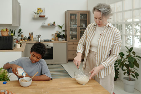 Woman whisking pancake batter while young boy pouring milk into cereal in cozy kitchen setting....