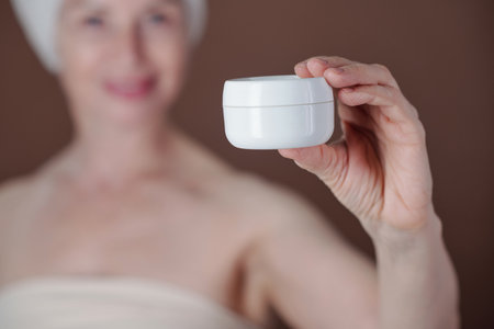 Close-up of woman showing a white cream jar while smiling against brown background. Demonstrating...