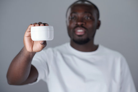 African American man in focus holding white plastic container with soft smile. Background blurred for emphasis on cream jar, likely meant for demonstration or reviewの写真素材