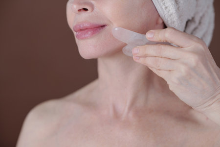 Close-up of woman massaging her cheek with quartz gua sha tool, rejuvenating skin. Brown background highlighting skincare practiceの写真素材