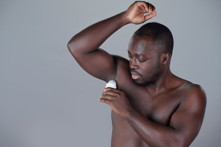 Black man standing shirtless and using deodorant under arm against grey background, showcasing grooming and hygiene habits with focus on arm position and product useの写真素材
