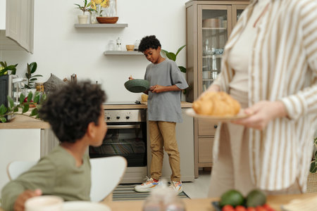 Morning scene of two African American children helping in kitchen while preparing breakfast with adult overseeing. Modern kitchen interior with green plants and wooden elementsの写真素材